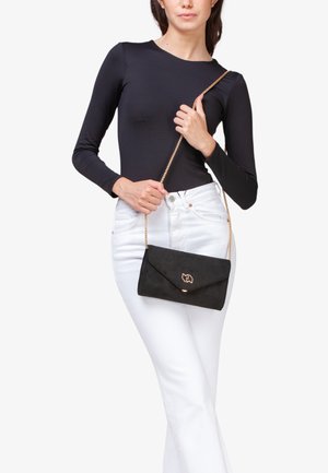 Bolso cruzado de ante negro con un cierre y emblema en tono dorado, con un diseño plano y correa de cadena, sostenido por una persona que lleva una blusa negra y pantalones blancos.