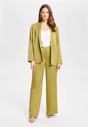 Femme debout portant un pantalon large vert olive et un blazer ouvert assorti sur un top blanc avec des talons ouverts et des boucles d’oreilles dorées.