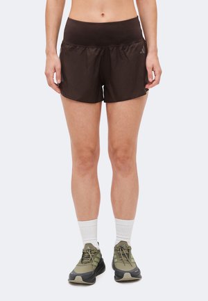 Άτομο που φοράει μαύρο αθλητικό σορτς, λευκές κάλτσες μέχρι τον αστράγαλο και πράσινα-μαύρα αθλητικά παπούτσια, στέκεται μπροστά σε απλό φόντο.