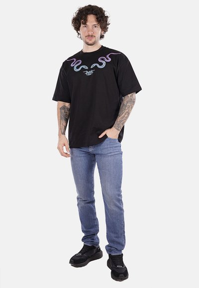 T-shirt nera in cotone con motivi di serpenti multicolori sul colletto, abbinata a jeans blu e sneaker nere.
