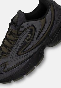 Svart och olivgrön sneaker med texturerade ytor, intrikat sömnad och en tjock, formad sula. Har en framträdande logotyp på sidan.