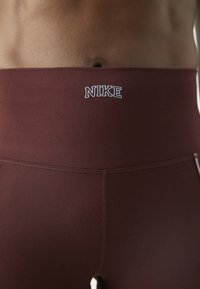 Burgundowe legginsy z wysokim stanem wykonane z elastycznego materiału o gładkiej fakturze. Posiadają białe logo "NIKE" na pasku.