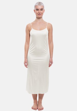 Femme souriante aux cheveux blancs courts portant une robe nuisette en satin blanc à fines bretelles, debout pieds nus sur un fond uni.