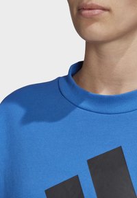 Sweat-shirt bleu avec un col rond côtelé, un tissu texturé et un grand logo noir sur le devant. Gros plan sur l'encolure et le haut de la poitrine.