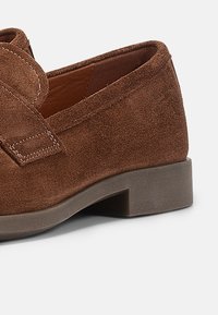 Brūnas zamša slip-on kurpes ar platu gumijas zoli, ar izšūtu siksniņas detaļu. Tuvplāns, kas izceļ tekstūru un dizainu.
