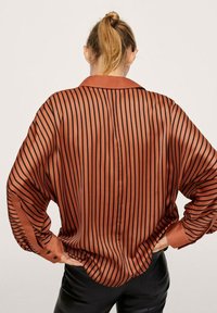 Camicia di seta con strisce nere verticali su fondo ruggine, dotata di colletto e maniche larghe con polsini.