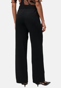 Pantalons larges noirs en tissu lisse, avec une taille élastique et deux poches arrière, associés à des escarpins noirs à bout pointu.