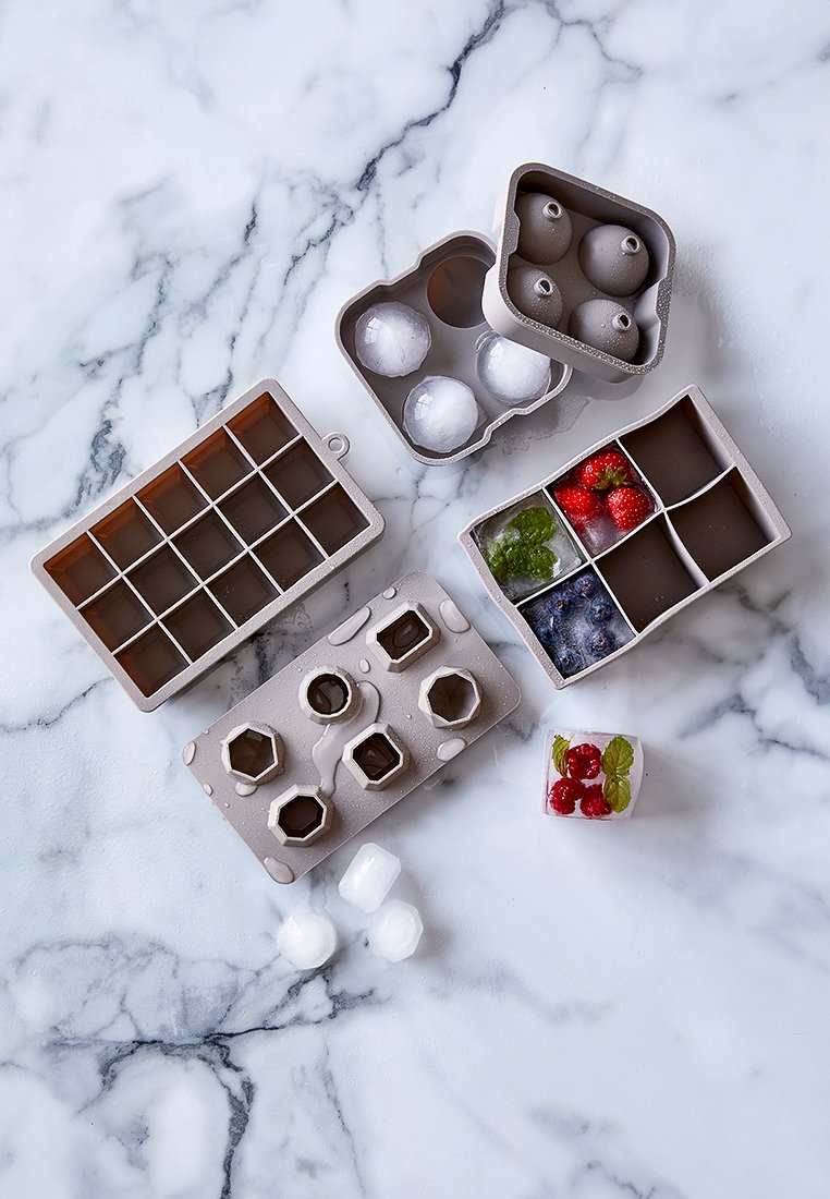Silicone-Eiswürfelschalen in Grau mit verschiedenen Formen: Kugeln, Sechsecke und rechteckige Abschnitte. Einige Würfel enthalten Obst und Kräuter.