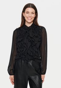 Black ruffled blouse with long sheer sleeves, high ruffled collar, front button closure, and textured detailing on fabric. Paired with black leather pants.