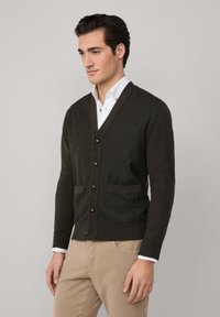 Muškarac u tamnozelenom kariranom kardiganu s dugim rukavima preko bijele košulje i bež hlačama, stoji naslonjen na svijetlu pozadinu.