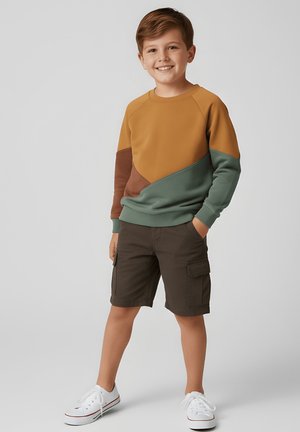 Uśmiechnięty chłopiec z rękami w kieszeniach, ubrany w kolorową bluzę, ciemne szorty typu cargo i białe trampki, stojący na jednolitym tle.