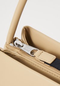 Bolso de mano de cuero beige con cierre de cremallera y herrajes plateados. Presenta un asa texturizada y un acabado suave en el cuerpo.