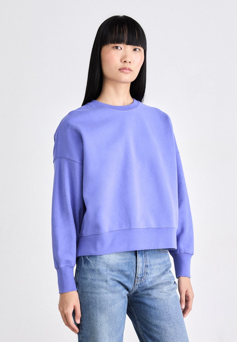 Sudadera morada recortada con cuello redondo y mangas largas, hecha de una tela suave, combinada con jeans de color azul claro.