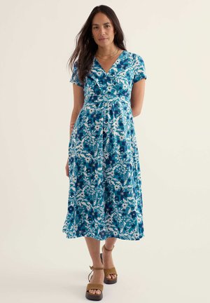 Naine seisab käed selja taga, seljas sinine lilleline midi kleit ja jalas pruunid platvorm-sandaalid, taust on ühtlane.