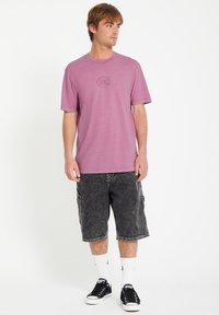 Lyse lilla kortærmet t-shirt med rund hals og præget logo på brystet, parret med mørke cargo-shorts og sorte sneakers.