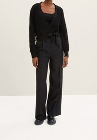 Černý pletený kardigan na zavázání v pase, v kombinaci s černými kalhotami s vysokým pasem a sklady, a masivními černými botami.