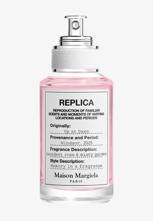 Flacon de parfum Maison Margiela Replica avec liquide rose, pulvérisateur argenté, étiquette indiquant le parfum "Up at Dawn" avec des notes de rose et de jardin embrumé.
