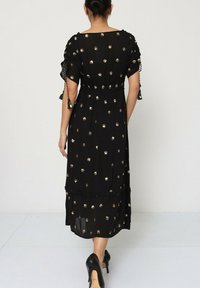 Robe maxi noire avec des embellissements en points dorés, des manches courtes et une taille cintrée. Le tissu semble léger avec une texture fluide.