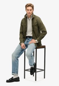 Chaqueta verde oliva sobre un suéter gris con cremallera, jeans azul claro y zapatos negros. El modelo está sentado en un taburete, con las manos apoyadas en el muslo.