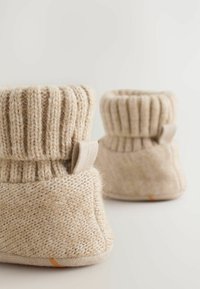 Rätknutna beiga tofflor med ribbade muddar, med en texturerad kropp och en liten tygslinga. Mjukt, mysigt material med en halkfri sula.