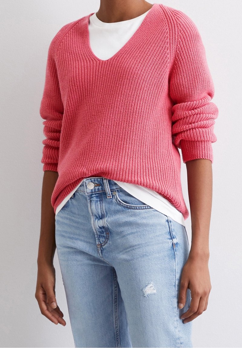 Pull en tricot rose corail avec un col en V et des manches retroussées, porté sur une chemise blanche. Associé à un jean bleu clair déchiré.