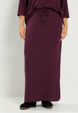 Donna che indossa una gonna lunga color viola scuro con coulisse in vita e scarpe nere slip-on, in piedi su uno sfondo semplice.