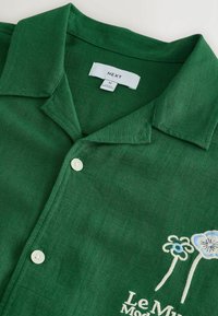 Chemise verte à col avec des boutons blancs, des fleurs bleues et blanches brodées, et le texte « Le Mur » près du bas droit du col.