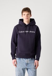 Tengerészkék kapucnis pulóver, zsinórral és elülső logóval, amelyen a "Calvin Klein Jeans" felirat olvasható. Puha anyagból készült, laza szabású, ribbelt szegéllyel.