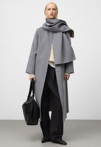 Grå ullkappa med draperad design, öppen front och knappar. Kombinerad med beige tröja, svarta vida byxor och en svart läderväska.