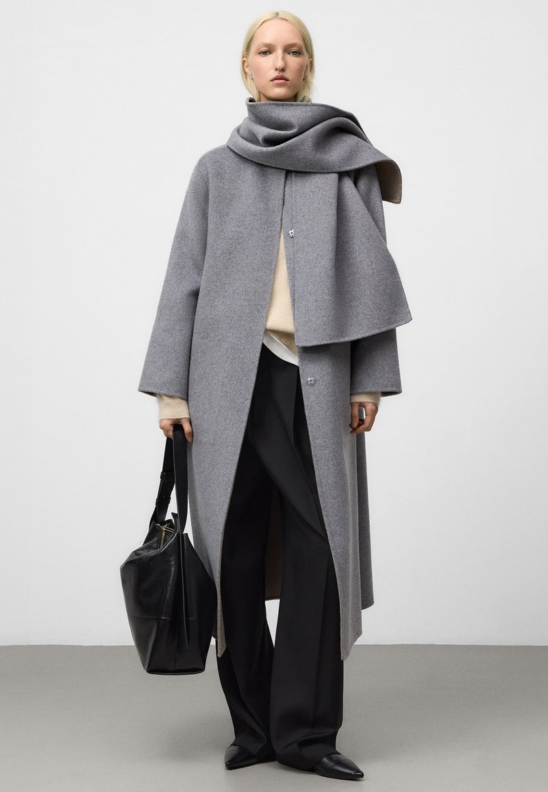 Grå ullkappa med draperad design, öppen front och knappar. Kombinerad med beige tröja, svarta vida byxor och en svart läderväska.
