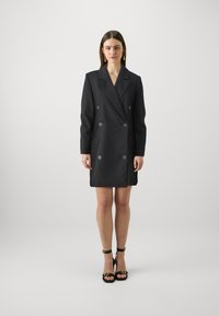 Rochie blazer neagră cu două rânduri de nasturi mari, guler cu colțuri, mâneci lungi și textură netedă, ajungând până la mijlocul coapsei.