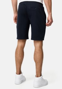 Marineblå shorts lavet af blødt stof, med en baglomme med knap, der når lige over knæet, parret med hvide sneakers.