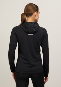 Giacca nera con cappuccio e finitura testurizzata. Presenta un design aderente e un logo sul retro. Maniche lunghe con accenti neri solidi.
