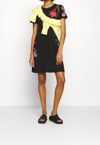 Schwarzes Kleid mit floraler Stickerei in Pink und Grün. Gelbes Strickoberteil ist über die Schultern drapiert. Schwarze Sandalen mit flacher Sohle.