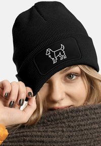 Black knit beanie with a textured surface and a rectangular black patch featuring a white dog silhouette. Close-up of a hand and face.