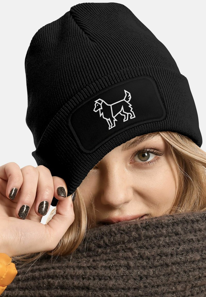 Black knit beanie with a textured surface and a rectangular black patch featuring a white dog silhouette. Close-up of a hand and face.