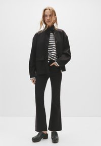 Mladá žena stojí před bílým pozadím, oblékla si černou bundu, pruhovanou dlouhou rukávu košili, černé rozšířené džíny a černé dřeváky.