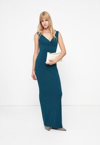Teal színű, testhez álló maxi ruha mély V-kivágással és ujjatlan kivitelben. A modell egy fehér táskát tart és neutrális színű magassarkút visel.