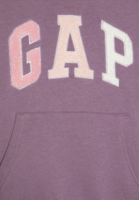 Sudadera morada con un gran bordado suave de la palabra "GAP" en rosa claro y blanco. Incluye un bolsillo frontal y una textura suave.