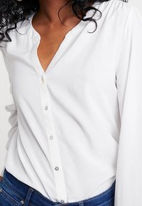 Chemise blanche boutonnée avec un col en V, manches longues et texture subtile. Dotée de boutons transparents sur le devant, associée à un jean en denim.