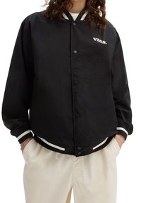 Osoba ubrana w czarną kurtkę varsity Vans z białymi paskami na mankietach i u dołu, w połączeniu z kremowymi spodniami, z rękami w kieszeniach.