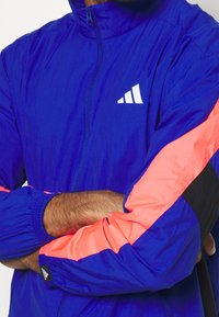 Rompevientos azul ligero con cremallera completa, con mangas vibrantes en naranja y negro. Incluye un logo blanco en el pecho.