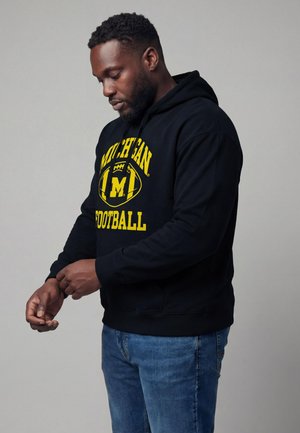 Mann trägt einen schwarzen Michigan Football-Hoodie und richtet seinen Ärmel, stehend vor einem einfarbigen grauen Hintergrund.
