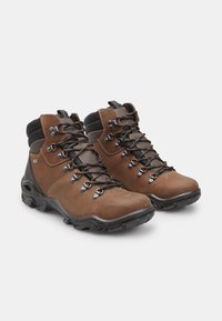 Barna bőr túrabotók fekete textilkollárral, fém fűzőlyukakkal és fekete gumi talppal, párnázott bokarészes támogatással.