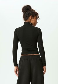 Femme avec des cheveux bouclés en chignon, portant de grandes boucles d'oreilles créoles, un turtleneck noir ajusté à manches longues et un pantalon noir ample, de dos.