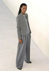 Graue weitgeschnittene Hose kombiniert mit einem gemusterten schwarz-weißen Blazer. Das Modell trägt ein graues Oberteil und auffällige Ohrringe und steht vor einem hellen Hintergrund.