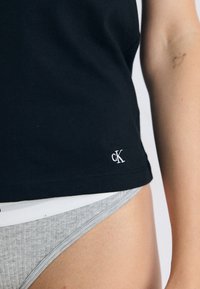 Un gros plan du bas du torse d'une personne portant un t-shirt noir avec un petit logo "CK", associé à des sous-vêtements gris. L'arrière-plan est neutre.