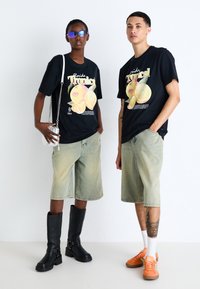T-shirts graphiques noirs avec illustration de fruits, shorts en denim verts, bottes noires jusqu'aux genoux et baskets orange présentent des textures et des couleurs contrastées.