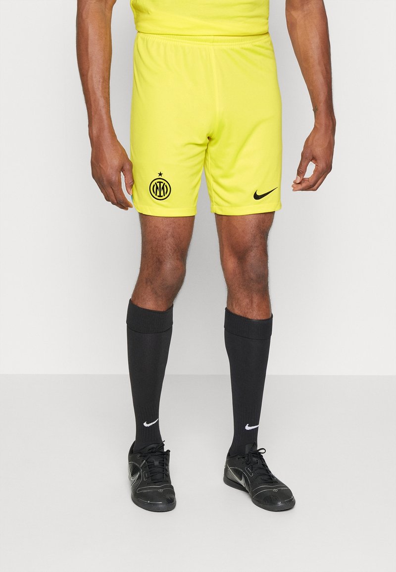 Shorts de football Nike jaunes avec logo noir, associés à des chaussettes noires montantes et des chaussures de sport noires. Le tissu semble léger et doux.