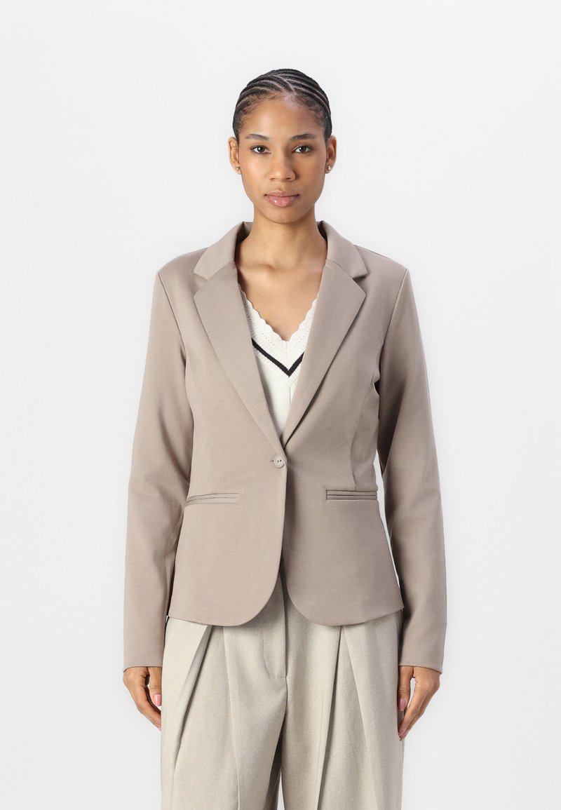Světle hnědý elegantní blazer s klopou, zapínáním na jeden knoflík, dvěma předními kapsami a hladkým materiálem. V kombinaci s krémovým topem a světlými kalhotami.